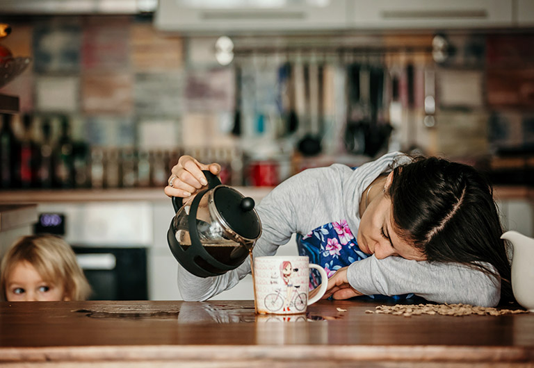 Tired mother, trying to pour coffee in the morning. Woman lying on kitchen table after sleepless night, trying to drink coffee